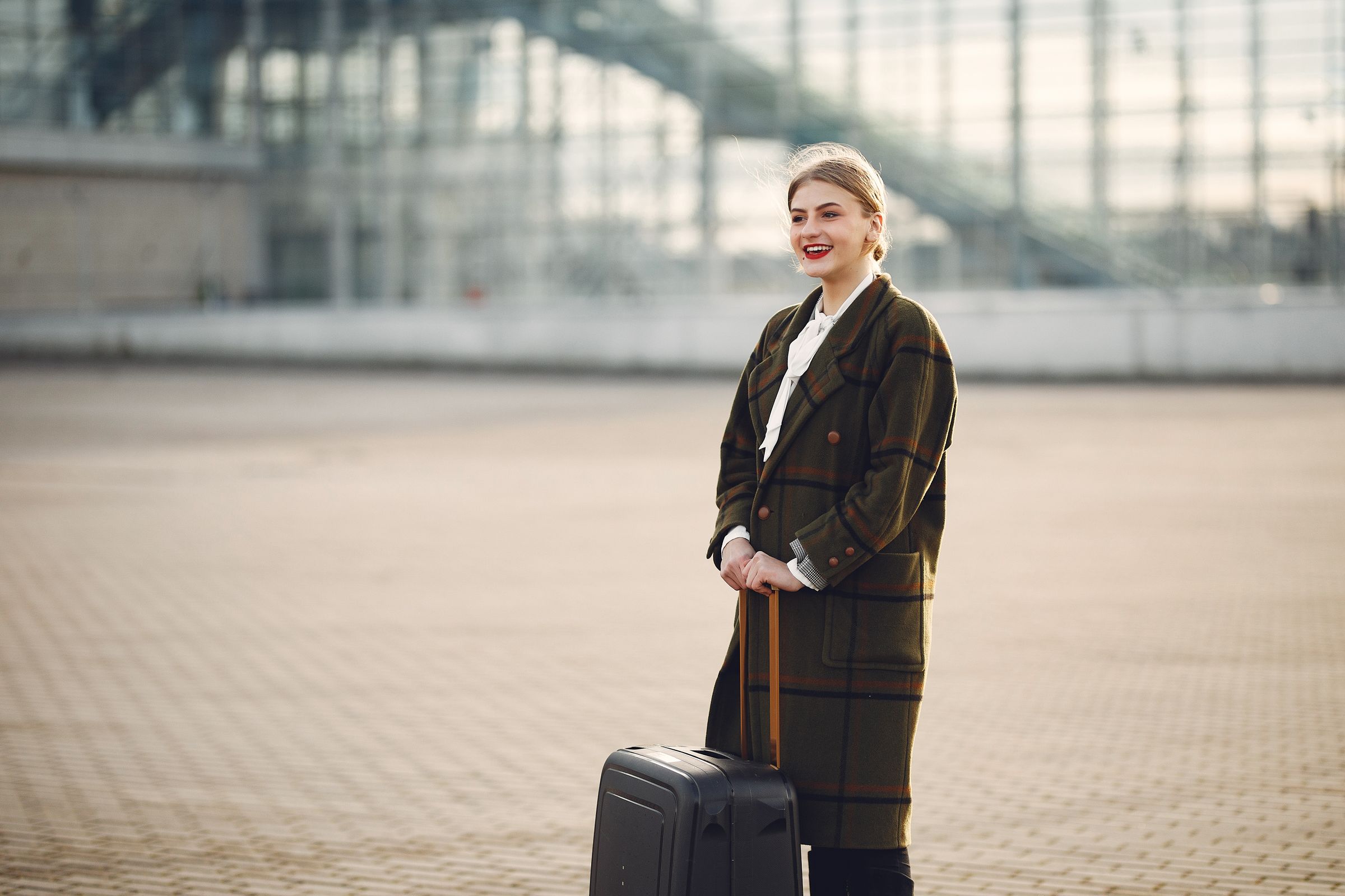A young woman smiling with her suitcase outside an airport, ready to travel abroad